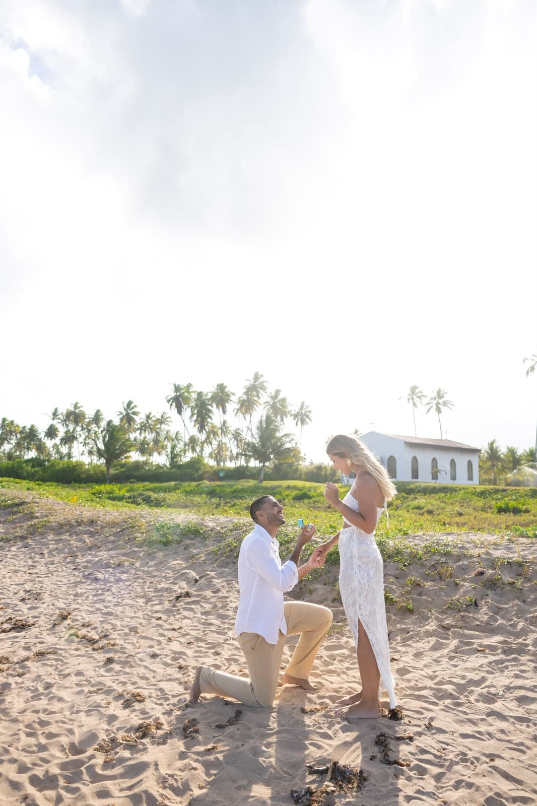 Victor ajoelhado na areia pedindo Camila em casamento, com a capela ao fundo