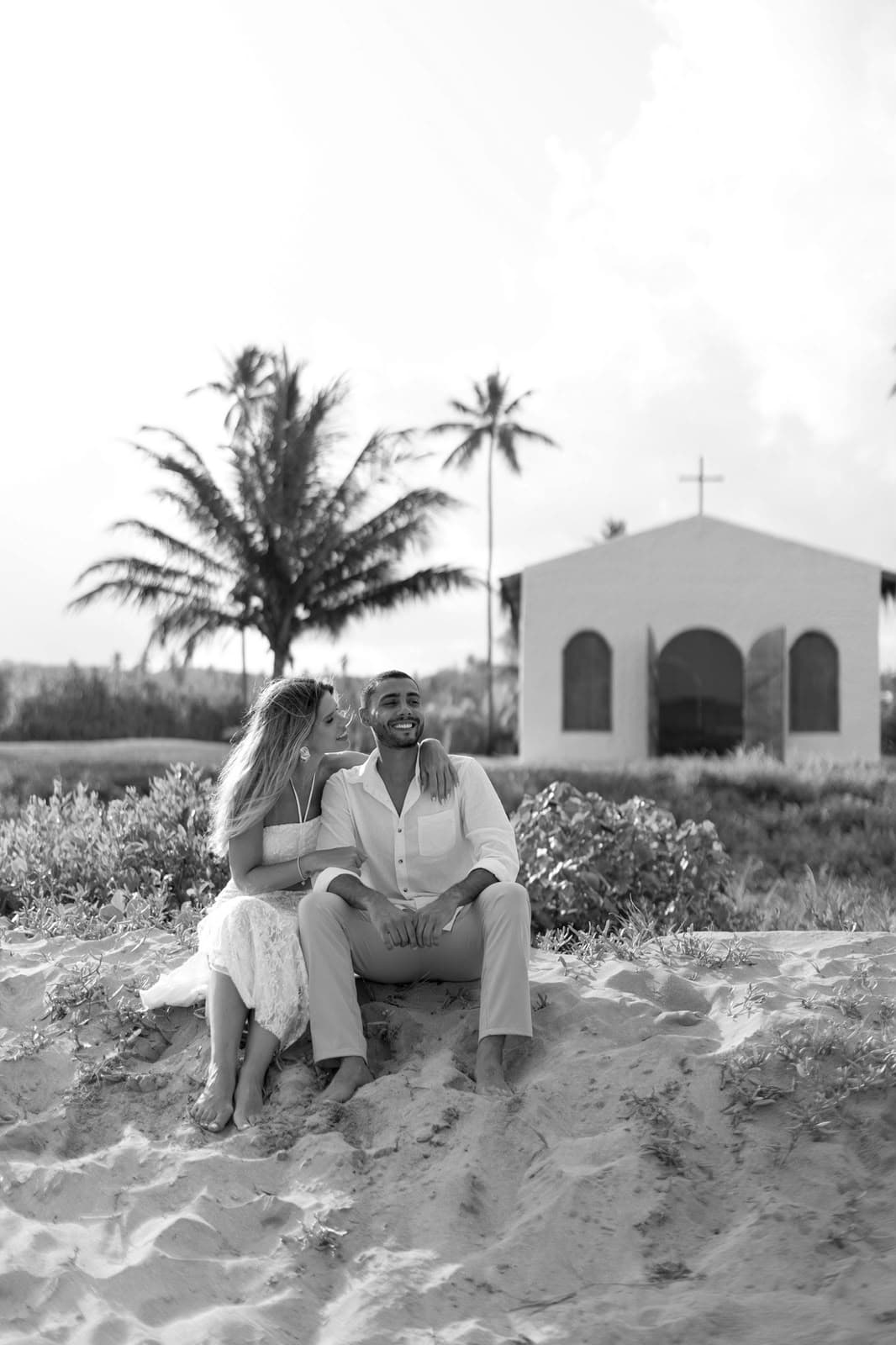 Casal sentado na areia em frente à capela, em preto e branco